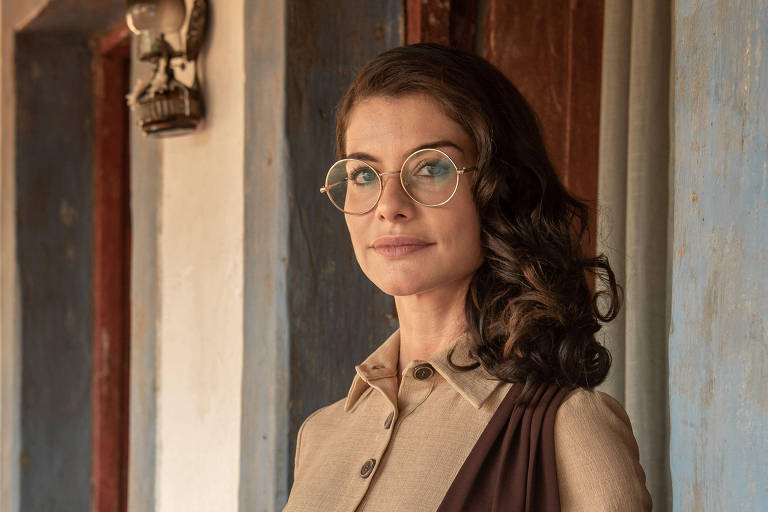 A imagem mostra uma mulher em pé ao lado de uma parede de uma casa com detalhes rústicos. Ela está usando uma roupa de cor clara com um detalhe marrom e tem cabelo ondulado. A mulher está olhando para a câmera e usa óculos. Ao fundo, há uma janela e uma planta visível.