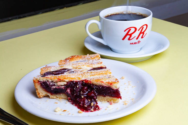 Na imagem, há um prato com uma fatia de sobremesa, que parece ser um bolo ou torta com recheio de frutas vermelhas, coberto com açúcar de confeiteiro. Ao lado, há uma xícara de café fumegante em um pires, com a letra 'RR' estampada na frente. O fundo é escuro e a mesa é de cor amarela.