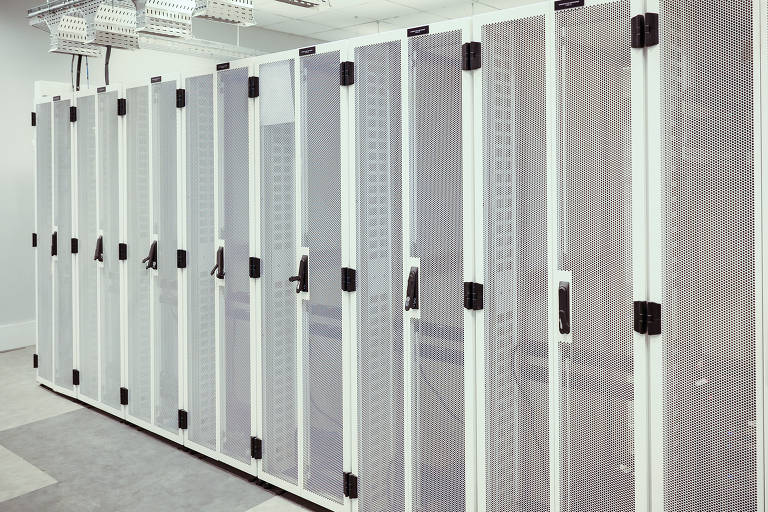 A imagem mostra uma sala de servidores com várias unidades de armazenamento dispostas em fileiras. As unidades possuem portas de malha metálica e são organizadas em um ambiente limpo e bem iluminado. O piso é de cor clara e há prateleiras suspensas no teto.