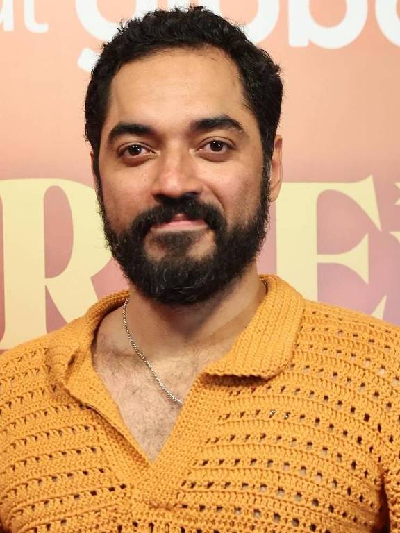 Um homem posando em um evento, vestindo uma camisa de crochê laranja e calças brancas. Ele tem barba e cabelo escuro, e está sorrindo para a câmera. Ao fundo, há um painel com o texto 'GUERREIRO DO SOL' e o logotipo da plataforma de streaming.