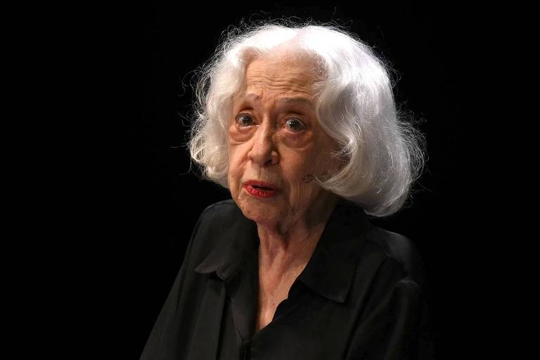 A imagem mostra uma mulher idosa com cabelo branco e ondulado, usando uma blusa preta. Ela está olhando para a câmera com uma expressão séria, em um fundo escuro que destaca seu rosto e cabelo. A iluminação é suave, criando um contraste com o fundo.