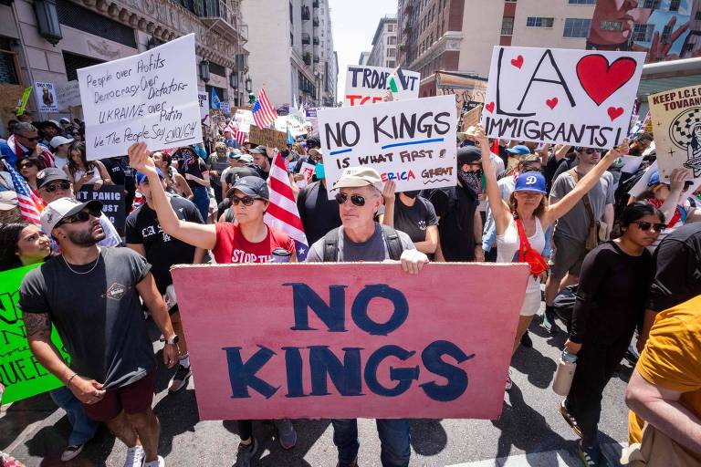 A imagem mostra um protesto em uma rua, com várias pessoas segurando cartazes e bandeiras. No centro, um grupo de manifestantes exibe um grande cartaz escrito 'NO KINGS'. Outros cartazes têm mensagens como 'LA ♥ IMMIGRANTS!' e 'TROVA IT TEA'. As pessoas estão vestidas de maneira casual e algumas usam óculos escuros. O ambiente é urbano, com prédios ao fundo.