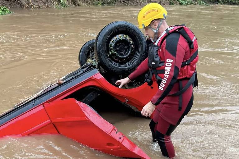 Un uomo con indosso una muta da sub e un casco giallo è in piedi nell'acqua di un fiume, accanto a un'auto rossa capovolta. L'auto è parzialmente sommersa, con le ruote rivolte verso l'alto, e l'acqua del fiume è di colore marrone. L'area circostante è ricoperta da una fitta vegetazione.