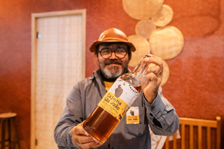 Um homem com barba e óculos, usando uma camisa listrada e um chapéu de couro, está sorrindo enquanto segura uma garrafa de bebida. A garrafa tem um rótulo que diz 'Cachaça São João'. Ao fundo, há uma parede decorada com elementos de palha e uma porta de madeira.