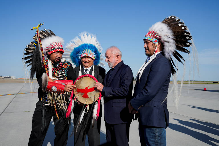 A imagem mostra quatro homens em um ambiente ao ar livre, possivelmente em um aeroporto. Dois deles estão vestidos com trajes tradicionais indígenas, incluindo cocares coloridos e penas. Um dos homens está segurando um tambor, enquanto outro parece estar em uma conversa. O céu está claro e azul, e o chão é de concreto.