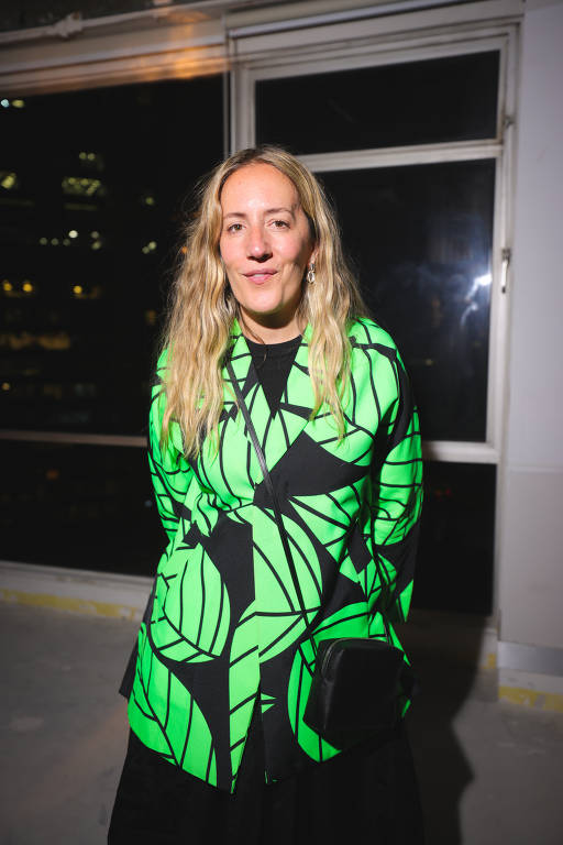 Uma mulher com cabelo longo e liso, usando um casaco verde neon com estampas de folhas. Ela está em pé em um ambiente interno, com janelas ao fundo mostrando luzes da cidade à noite. O chão parece ser de cimento e a iluminação é suave.