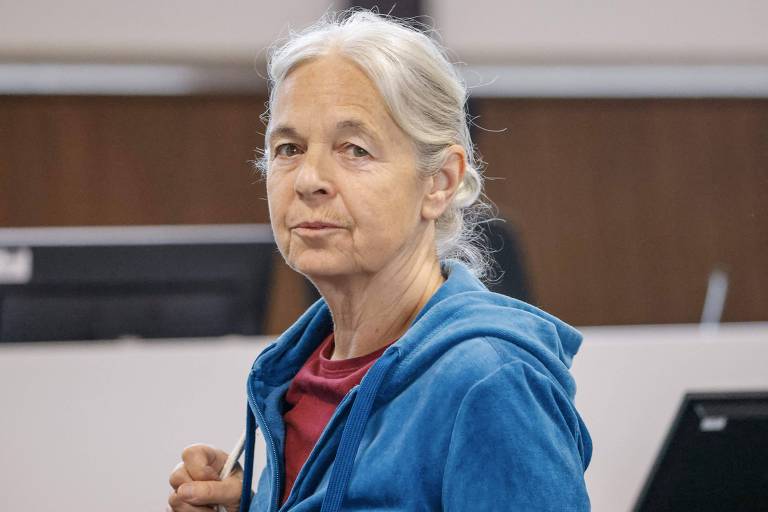 A imagem mostra uma mulher de cabelos grisalhos, usando um moletom azul e uma camiseta vermelha. Ela está em um ambiente interno, com mesas e computadores ao fundo. A mulher está olhando para a câmera com uma expressão neutra.