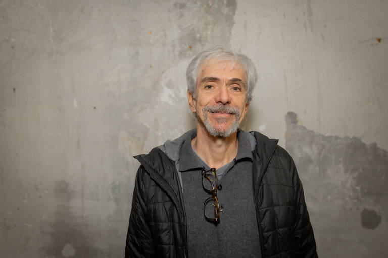 Um homem de cabelos grisalhos e barba curta, sorrindo para a câmera. Ele está usando uma jaqueta preta e uma camiseta cinza. Ao fundo, há uma parede de concreto com manchas e texturas visíveis.