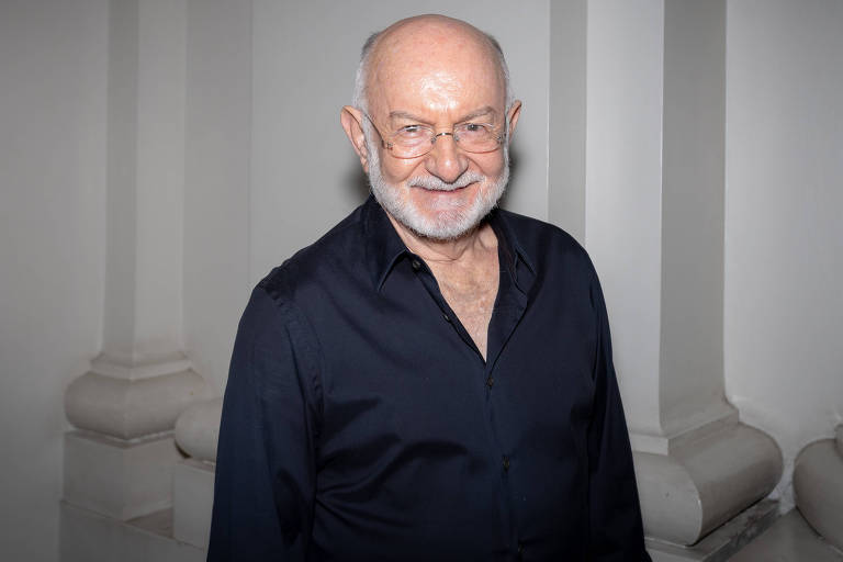 Um homem idoso com cabelo grisalho e barba branca, usando óculos e uma camisa preta, está sorrindo em um ambiente com paredes claras e colunas. Ele parece estar posando para a foto.