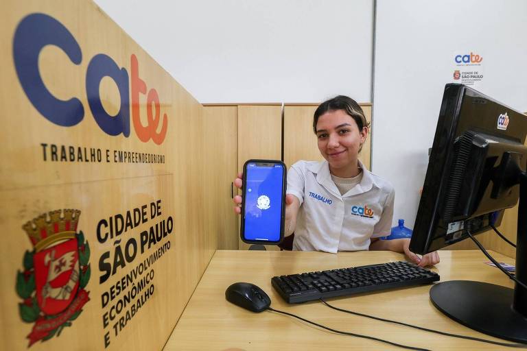 Uma funcionária do CATe (Centro de Apoio ao Trabalho e Empreendedorismo) de São Paulo está sorrindo e segurando um celular com a tela visível. Ao fundo, há uma mesa com um computador e um mouse, além de uma placa com o nome e logotipo do CATe, que inclui a frase 'TRABALHO E EMPREENDEDORISMO'. A placa também exibe o brasão da cidade de São Paulo e informações sobre o desenvolvimento econômico e trabalho.