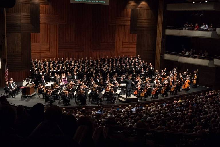 A imagem mostra um concerto em um auditório, onde uma orquestra e um coral estão se apresentando. A orquestra está posicionada na parte inferior da imagem, com músicos tocando instrumentos como violinos, violoncelos e percussão. Atrás deles, há um grande coral de cantores vestidos de preto. O auditório está cheio de espectadores, e a iluminação destaca os músicos e o coral. Ao fundo, há painéis de madeira que decoram a parede do auditório.