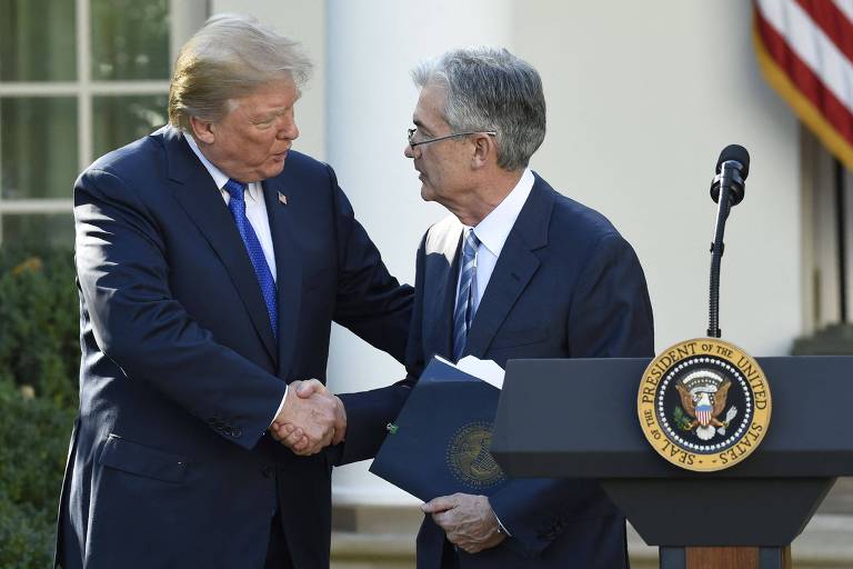Na imagem, duas pessoas estão se cumprimentando em um ambiente ao ar livre, possivelmente no jardim da Casa Branca. Uma delas está vestida com um terno escuro e uma gravata azul, enquanto a outra usa um terno claro. Ao fundo, é possível ver uma bandeira dos Estados Unidos e um púlpito com o selo presidencial. A cena parece ser de um evento formal.