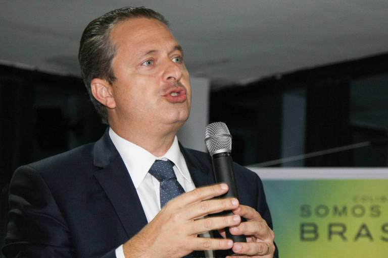 Um homem em um terno escuro está falando ao microfone. Ele gesticula com as mãos enquanto se dirige a uma audiência. Ao fundo, há um banner com a frase "SOMOS TODOS BRASÍLIA" e imagens de outras pessoas. O ambiente parece ser um evento político.
