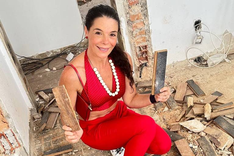 Uma mulher está sentada no chão de um ambiente em reforma, cercada por tábuas de madeira e detritos. Ela usa um conjunto vermelho e segura duas tábuas de madeira nas mãos. O ambiente ao fundo mostra paredes expostas e fios elétricos visíveis.