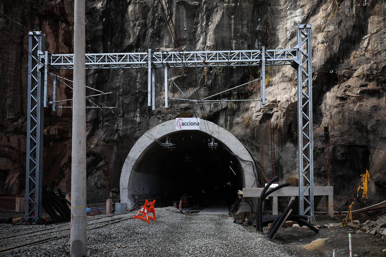 A imagem mostra a entrada de um túnel em construção, com uma estrutura metálica ao redor da abertura. O túnel é grande e escuro, e há uma placa com a palavra 'Acciona' visível na parte superior. Na frente do túnel, há uma barreira laranja e alguns trilhos de trem visíveis no chão, cercados por pedras. As paredes do túnel são de rocha exposta, e a iluminação é escassa, criando um ambiente sombrio.
