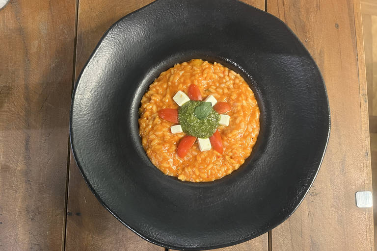 Um prato de risoto laranja, decorado com cubos brancos de queijo e pedaços de tomate, com um centro de pesto verde. O risoto está servido em um prato preto, com uma borda larga, sobre uma mesa de madeira.