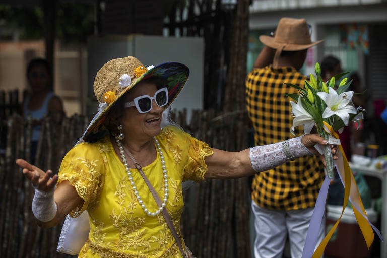 Una donna anziana balla e si diverte a una festa. Indossa un abito giallo, un grande cappello decorato con fiori e occhiali da sole bianchi. In una mano tiene un mazzo di fiori bianchi con nastri colorati. Sullo sfondo, una persona con una camicia a quadri gialli e neri e una staccionata di legno. L'ambientazione sembra quella di una festa all'aperto.