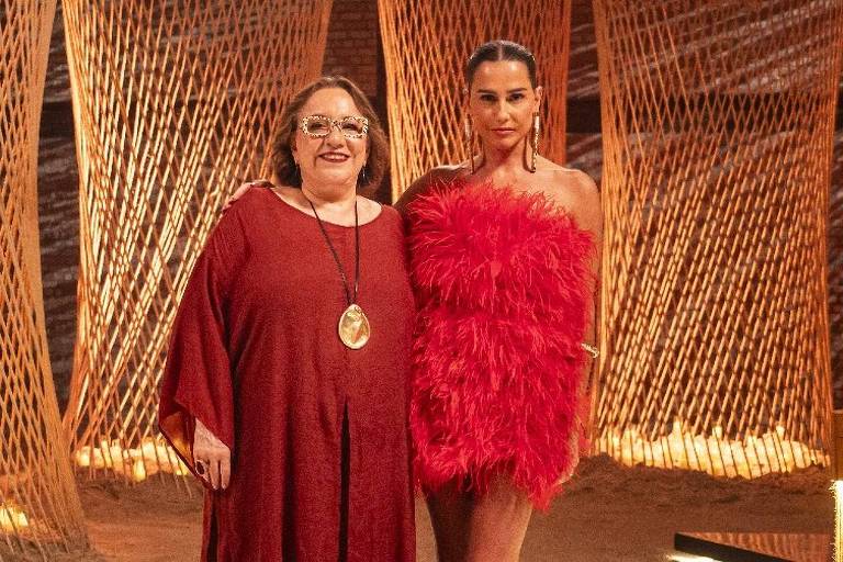 A imagem mostra duas mulheres em um ambiente decorado com estruturas de madeira entrelaçadas e iluminação suave. A mulher à esquerda está vestindo uma roupa longa e vermelha, com um colar grande. A mulher à direita usa um vestido curto e vibrante, com detalhes de penas, e está em pose confiante. O chão é de areia e há uma iluminação em LED ao longo do caminho.
