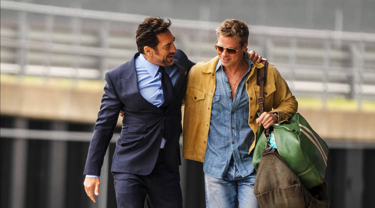 Dois homens estão caminhando lado a lado em um ambiente urbano. Um deles está vestido com um terno escuro e uma camisa azul, enquanto o outro usa uma camisa jeans e uma jaqueta amarela. O homem da jaqueta carrega uma bolsa grande e parece estar sorrindo. Eles parecem estar se divertindo e conversando, com um dos homens colocando o braço em volta do outro.