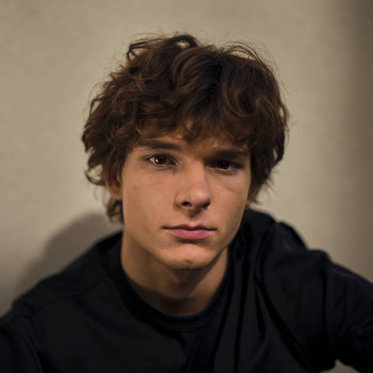A imagem mostra um jovem com cabelo cacheado e bagunçado, olhando diretamente para a câmera. Ele está vestindo uma camiseta preta e parece estar em um ambiente com iluminação suave, que destaca seu rosto. O fundo é de uma cor neutra, o que faz com que o foco permaneça no jovem.