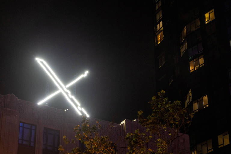 A imagem mostra um letreiro em forma de 'X' iluminado por luzes brancas, situado no topo de um edifício. O fundo é escuro, indicando que a foto foi tirada à noite. À direita, há um prédio com janelas iluminadas, sugerindo que algumas pessoas estão dentro. A vegetação na parte inferior da imagem é visível, com algumas folhas e galhos.