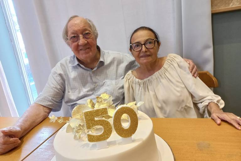 Um casal está sentado em uma mesa, sorrindo para a câmera. Eles estão ao lado de um bolo de aniversário decorado com flores e com o número 50 em destaque. O homem está usando uma camisa clara e a mulher está vestindo uma blusa branca. O ambiente parece ser bem iluminado, com uma cortina clara ao fundo.
