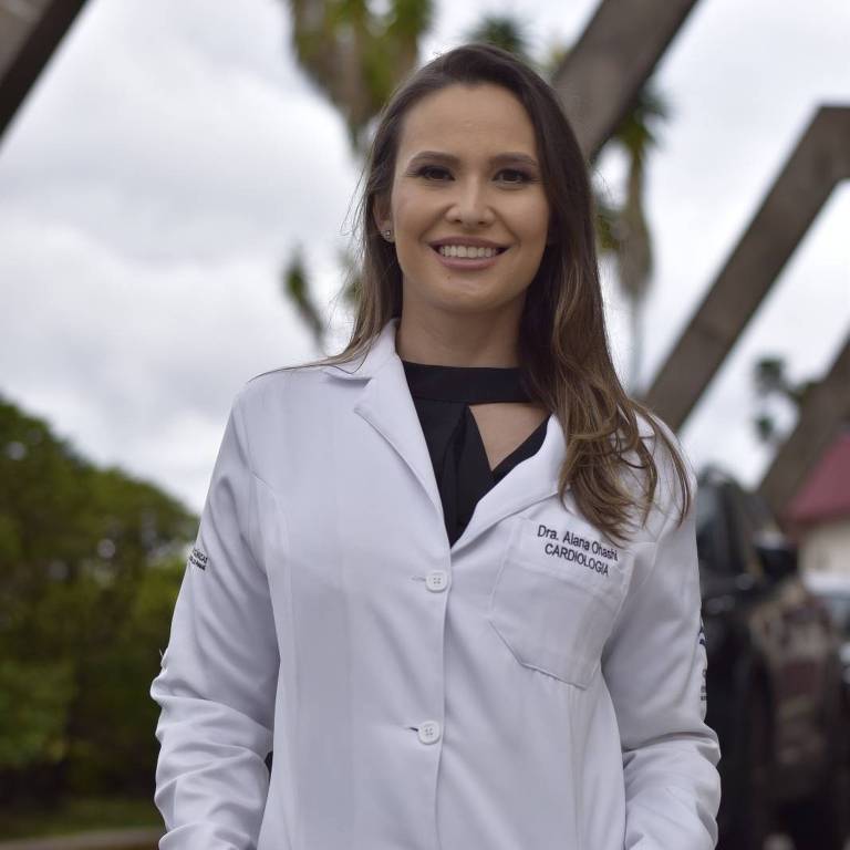 Uma mulher está em pé, usando um jaleco branco, com as mãos nos bolsos. Ao fundo, há estruturas metálicas e árvores, sob um céu nublado. A mulher tem cabelo longo e liso, e está sorrindo.