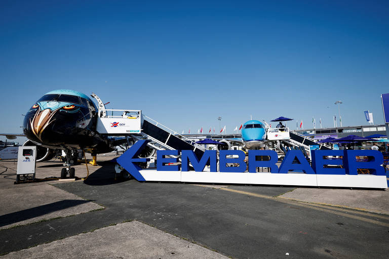 A imagem mostra um avião da Embraer em uma exposição ao ar livre, com um design de pintura que apresenta uma cabeça de ave. Em primeiro plano, há um grande letreiro azul com a palavra 'EMBRAER'. Ao fundo, é possível ver outro avião e um céu claro e ensolarado.