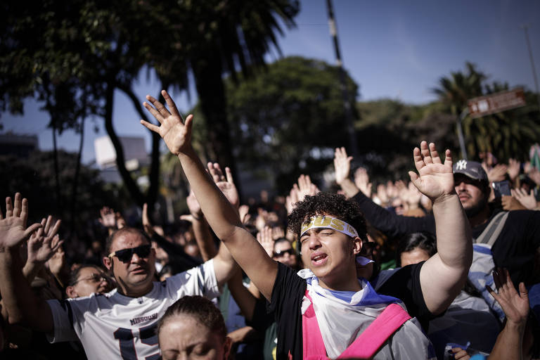 A imagem mostra uma multidão em uma manifestação ao ar livre, com várias pessoas levantando as mãos. No primeiro plano, um jovem com uma faixa na cabeça e uma camiseta colorida parece estar em um momento de emoção ou oração. Ao fundo, há outras pessoas com as mãos levantadas, e árvores e um céu claro são visíveis.