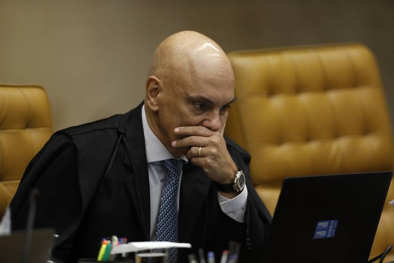 Foto do ministro Alexandre de Moraes no plenário do STF. O ministro, um homem branco, careca, veste uma toga preta sobre o terno. Ele está sentado em uma poltrona bege, com a mão esquerda sobre a boca enquanto olha para a tela de um laptop