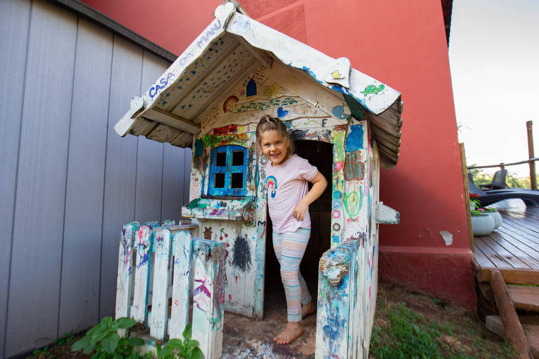 Uma criança está saindo de uma casinha de brinquedo colorida, que apresenta várias pinturas e desenhos nas paredes. A criança está sorrindo e usa uma camiseta clara e leggings listradas. A casinha tem uma porta e janelas azuis, e está situada em um ambiente externo com grama ao redor. Ao fundo, há uma parede vermelha e uma área de deck de madeira.