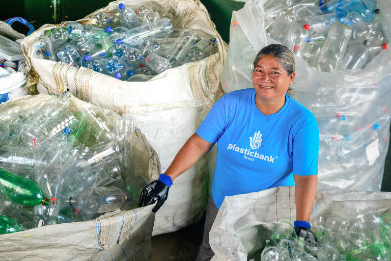 Uma mulher sorridente está em um ambiente de reciclagem, cercada por sacos grandes cheios de garrafas plásticas. Ela usa uma camiseta azul com a inscrição 'plasticbank' e luvas pretas. O fundo é composto por sacos brancos e garrafas plásticas de diferentes cores, indicando um esforço de reciclagem.