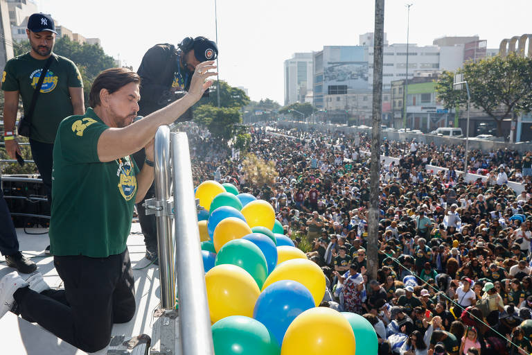 A imagem mostra uma grande multidão reunida em uma manifestação. No primeiro plano, uma pessoa de joelhos em um palco acena para a multidão. Ela está vestindo uma camiseta verde com um símbolo, e há outras pessoas ao fundo, algumas segurando câmeras. O cenário é urbano, com prédios ao fundo e balões nas cores verde, amarelo e azul, típicas da bandeira do Brasil. A atmosfera parece ser de celebração ou protesto.