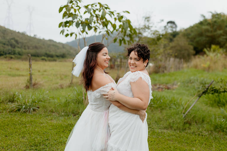A imagem mostra duas mulheres abraçadas e sorridentes, vestidas de branco, em um ambiente arborizado