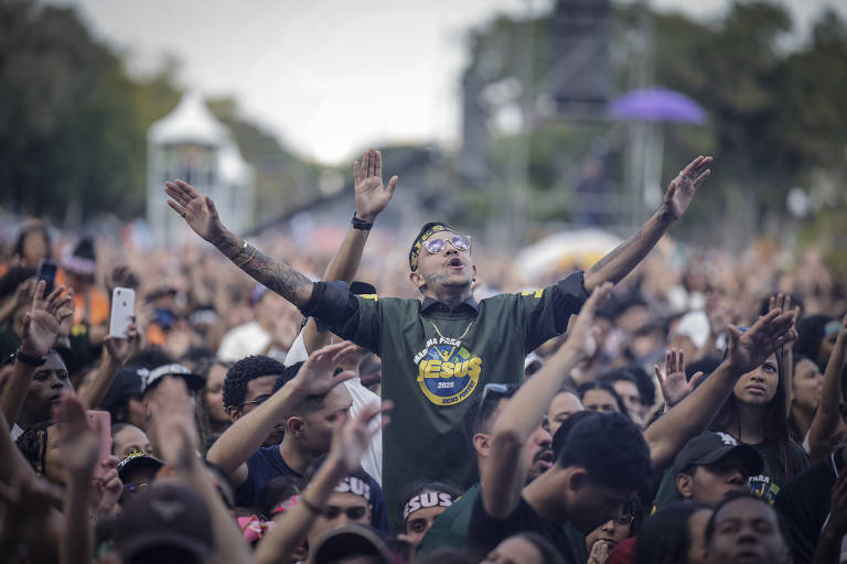 Uma grande multidão em um evento ao ar livre, com pessoas levantando as mãos e demonstrando entusiasmo. Um homem em destaque, usando uma camiseta preta com a inscrição 'SUS', está com os braços levantados e a cabeça inclinada para trás, expressando emoção. Ao fundo, outras pessoas também estão visíveis, algumas com os braços levantados, enquanto outras observam. O ambiente é festivo, com árvores e estruturas ao fundo.