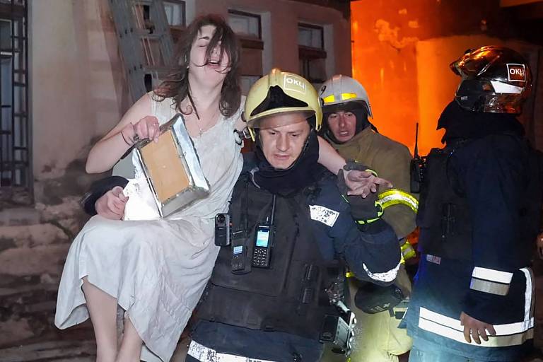 A imagem mostra um bombeiro carregando uma mulher vestida com um vestido claro, que parece estar em estado de choque. Ela segura uma bolsa e está sendo resgatada de um local que aparenta ter sofrido um incêndio. Ao fundo, há outros bombeiros e uma estrutura danificada, com luzes laranja refletindo na cena, indicando a presença de fogo. O ambiente é de emergência e tensão.