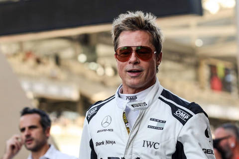 Cena de  F1: O Filme com Brad Pitt *** Local Caption *** Um homem vestido com um macacão de corrida branco e preto, com logotipos de patrocinadores visíveis, incluindo Mercedes e Pirelli. Ele usa óculos de sol e tem cabelo loiro. Ao fundo, há pessoas e uma pista de corrida, sugerindo um evento de automobilismo.