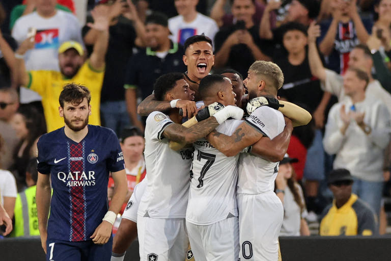 A imagem mostra um grupo de jogadores de futebol comemorando um gol, se abraçando em um momento de alegria. Ao fundo, há uma multidão de torcedores, alguns com expressões de entusiasmo. Em primeiro plano, um jogador com uniforme do Paris Saint-Germain parece desapontado, olhando para a cena de celebração. O ambiente é de um estádio, com bandeiras e cartazes visíveis entre os torcedores.