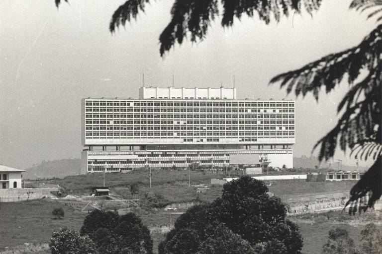 A imagem mostra um grande edifício moderno em estilo arquitetônico contemporâneo, com várias janelas e andares, situado em uma área verde. O edifício é predominantemente branco e possui uma estrutura retangular. Na parte inferior da imagem, há árvores e vegetação, enquanto o fundo é levemente nebuloso, sugerindo um dia nublado. O cenário é urbano, mas com uma sensação de tranquilidade devido à presença da natureza.