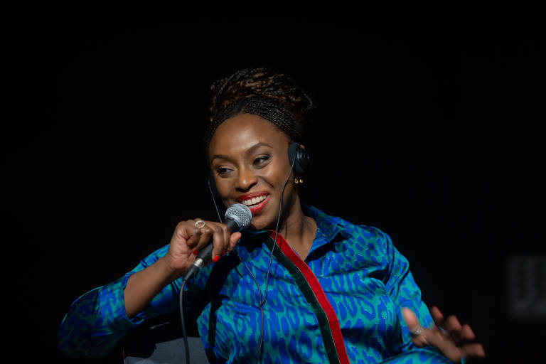 Uma mulher sorridente está falando em um microfone. Ela usa uma blusa azul com estampas e tem um fone de ouvido. O fundo é escuro, destacando sua expressão animada e envolvente.