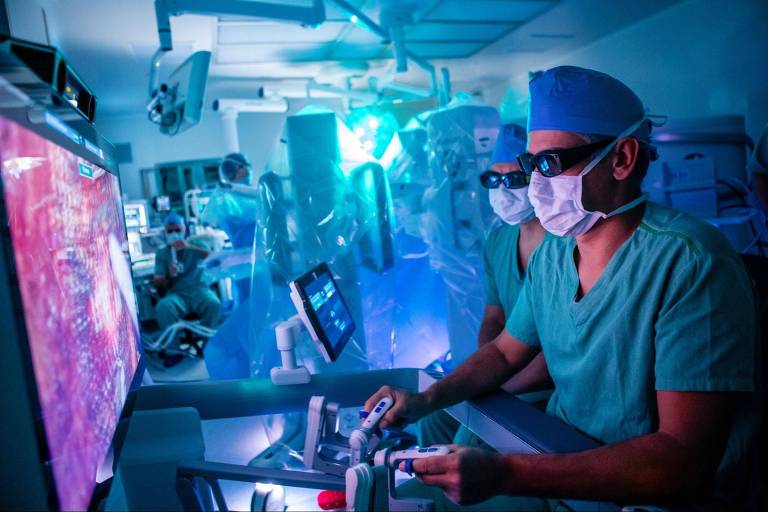 A imagem mostra uma sala de cirurgia moderna, onde dois cirurgiões estão utilizando um sistema robótico para realizar um procedimento. Eles estão vestidos com roupas cirúrgicas verdes e máscaras, e usam óculos especiais. Ao fundo, há uma tela grande exibindo imagens do procedimento, com iluminação azulada que cria um ambiente tecnológico. Outros profissionais de saúde podem ser vistos ao fundo, também envolvidos no processo.