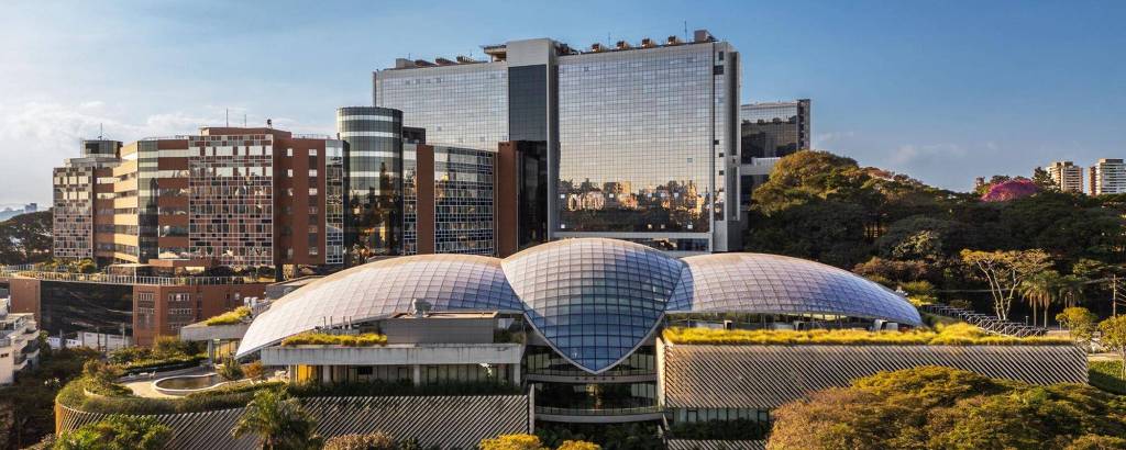 A imagem mostra um edifício moderno com uma estrutura curvilínea no primeiro plano, coberto por um telhado de vidro em forma de concha. Ao fundo, há vários prédios altos e modernos, refletindo a luz do sol. A vegetação ao redor é densa, com árvores e plantas, criando um contraste com a arquitetura urbana.