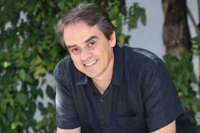 Um homem está sentado em um banco de madeira, sorrindo para a câmera. Ele usa uma camisa preta e calças azuis. Ao fundo, há uma vegetação verde, com folhas e galhos visíveis. A iluminação é natural, sugerindo um ambiente ao ar livre.