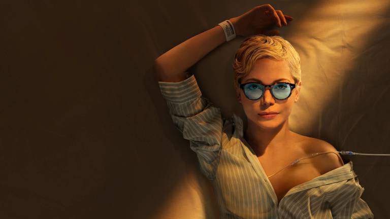 A imagem mostra uma mulher de cabelo curto e loiro, deitada em uma superfície clara. Ela usa uma camisa de botão com listras e óculos de sol azuis. A iluminação é suave, criando sombras e um ambiente tranquilo. Um fio é visível, sugerindo que ela pode estar em um ambiente hospitalar ou de recuperação.