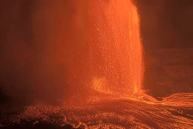 A imagem mostra uma erupção vulcânica com lava expelida em forma de jato, criando uma fonte de lava brilhante. O fundo é escuro, destacando o brilho intenso da lava que flui para baixo, formando um rio de lava.