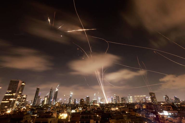 A imagem mostra uma vista noturna de uma cidade, onde arranha-céus iluminados se destacam contra o céu escuro. No céu, há várias trajetórias de luz, possivelmente de foguetes ou mísseis, que criam um efeito visual dinâmico. As nuvens estão presentes, e a cena transmite uma sensação de atividade intensa.