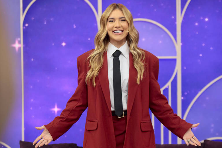 Uma mulher está em pé em um estúdio de televisão, usando um terno vermelho oversized com uma camisa branca e uma gravata preta. Ela está sorrindo e com os braços abertos, em frente a um sofá roxo. O fundo é decorado com elementos gráficos em tons de roxo e azul, com estrelas. O ambiente é bem iluminado e moderno.