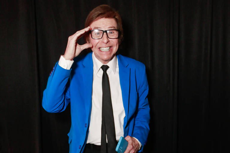 Um homem está posando para a foto em um fundo preto. Ele usa um paletó azul, uma camisa branca e uma gravata preta. O homem está sorrindo e fazendo um gesto de saudação com a mão. Ele também usa óculos grandes e segura um objeto na outra mão.