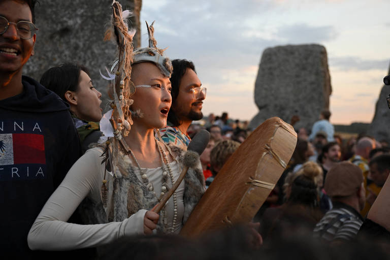 A imagem mostra um grupo de pessoas reunidas ao ar livre, com destaque para uma mulher no centro que está tocando um tambor. Ela usa um traje decorado com elementos naturais e tem um adereço na cabeça. Ao seu redor, há outras pessoas, algumas sorrindo e outras observando. O cenário é ao entardecer, com grandes pedras ao fundo, sugerindo um ambiente de celebração ou ritual.