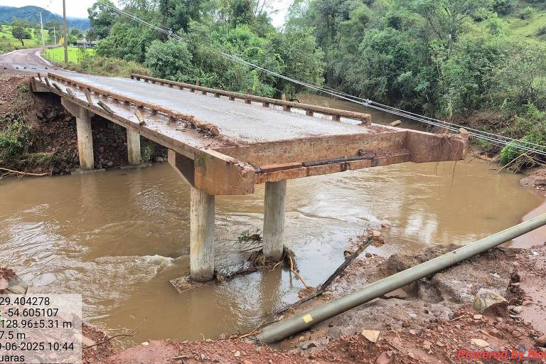 L'immagine mostra un ponte parzialmente danneggiato su un fiume. La struttura del ponte è sostenuta da piloni, ma sembra aver subito danni, con parte dell'impalcato mancante. Il fiume è pieno d'acqua e intorno ad esso si trova vegetazione, inclusi alberi e colline sullo sfondo. Nella parte inferiore dell'immagine è presente un tubo di drenaggio. Le informazioni tecniche sono visibili in basso a sinistra dell'immagine, tra cui latitudine, longitudine, altitudine e data.